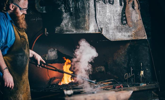 A blacksmith is dressed in a blue shirt and leather apron. He stands in front of a forge which is burning brightly. He holds a long metal rod that's j